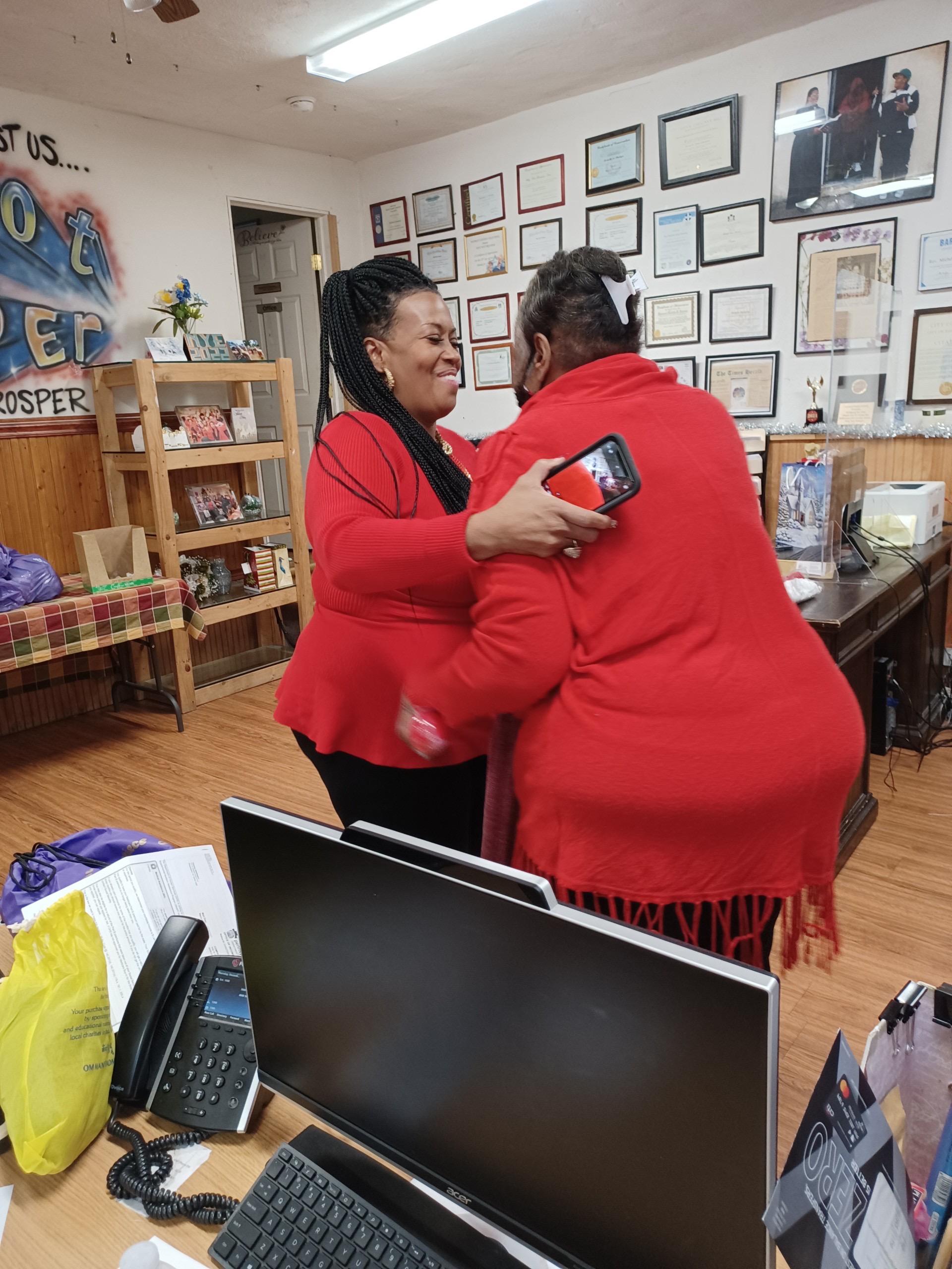 Rev. Michelle, wearing a red cardigan, hugs another woman in red at a Why Not Prosper facility.