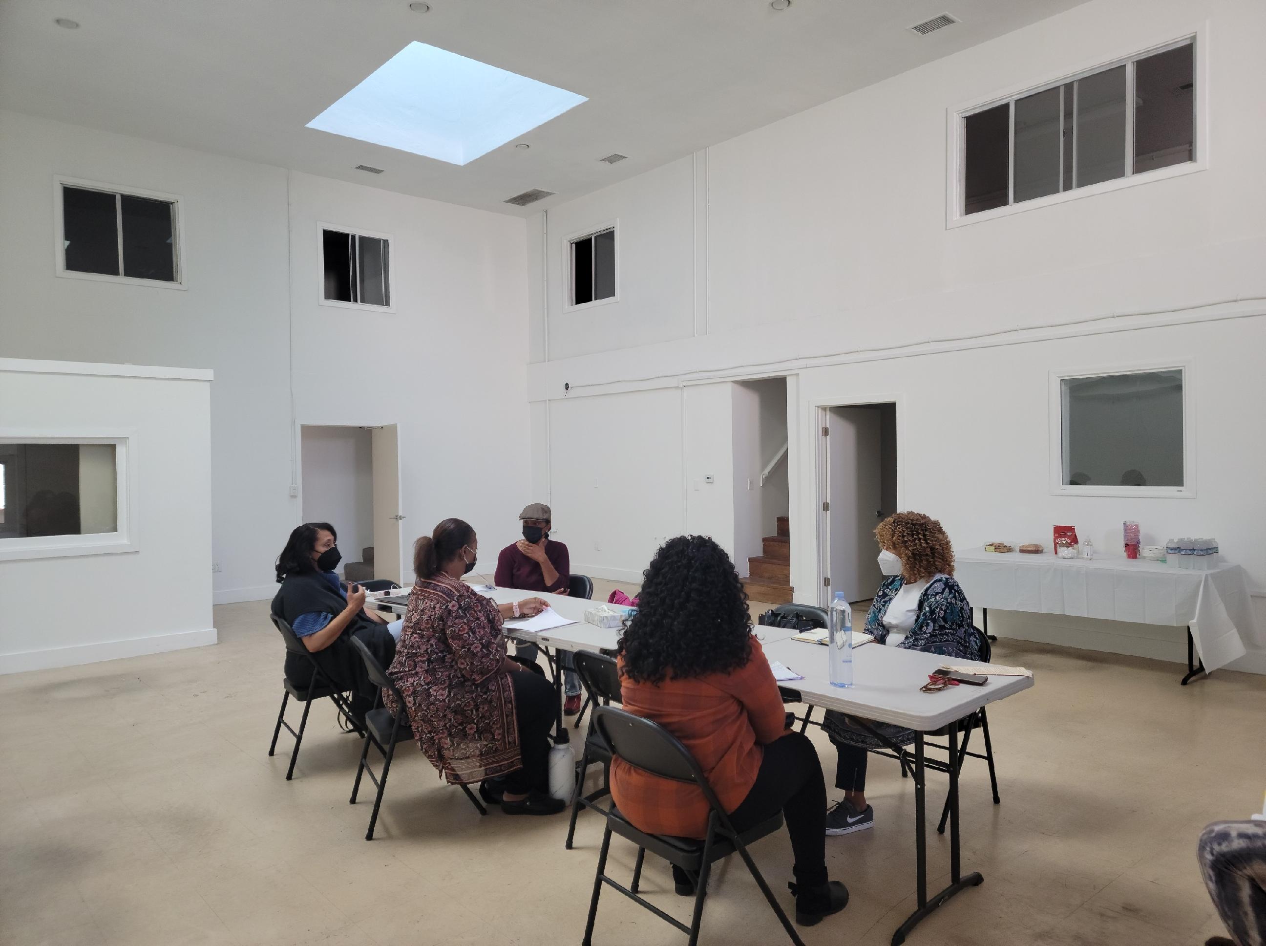 Five women in masks sit on around two folding tables in a large white room. Five women in masks sit on around two folding tables in a large white room.