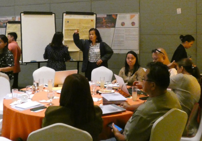 A woman standing in front of three large pieces of paper directs workshop participants seated at a round table.