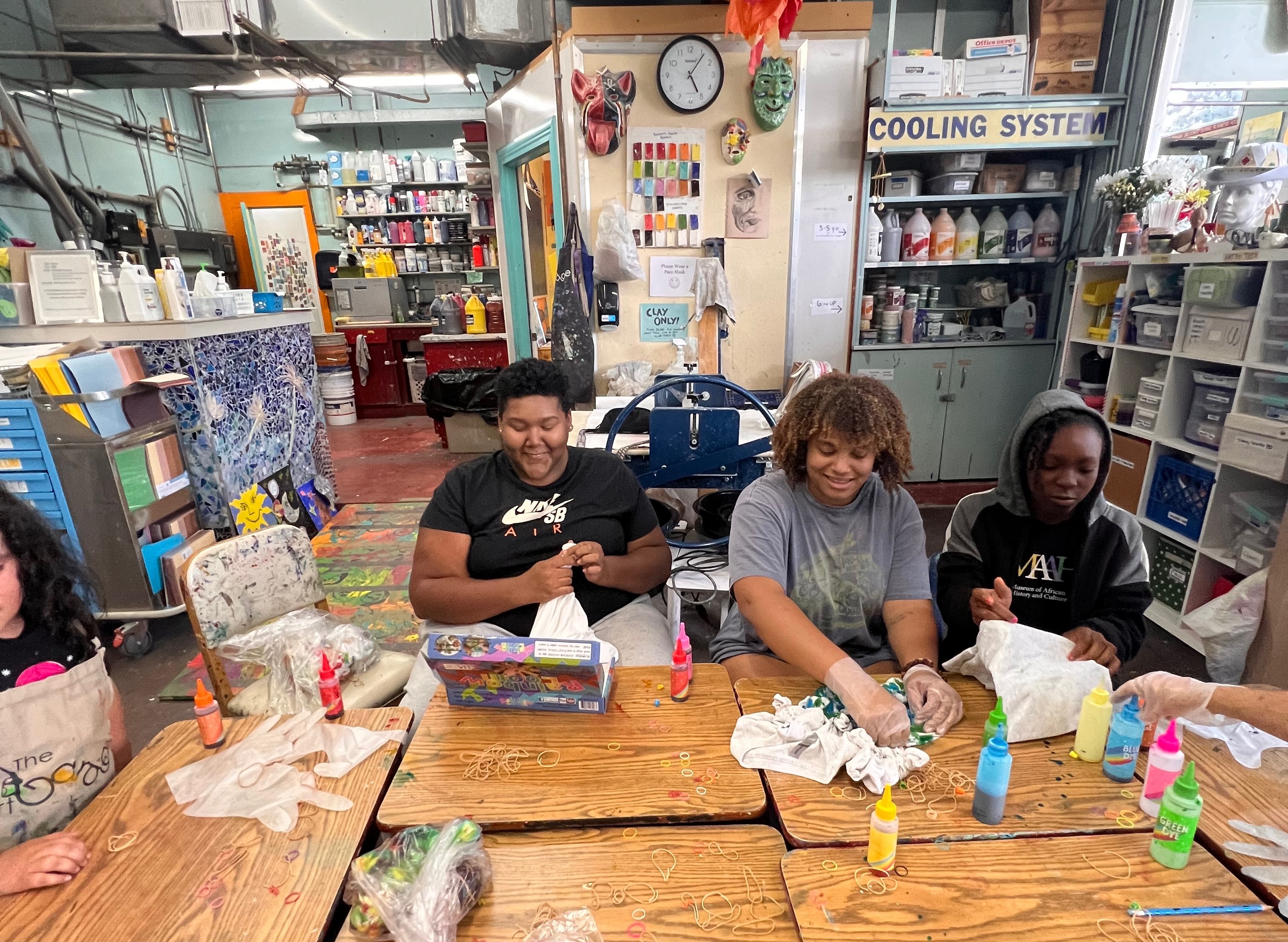 Three young people, seated at a desk, work with paints in a bustling art studio. Three young people, seated at a desk, work with paints in a bustling art studio.