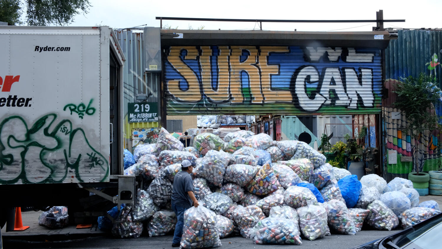 Photo of Sure We Can's sign behind a pile of recyclables in plastic bags. Photo of Sure We Can's sign behind a pile of recyclables in plastic bags.