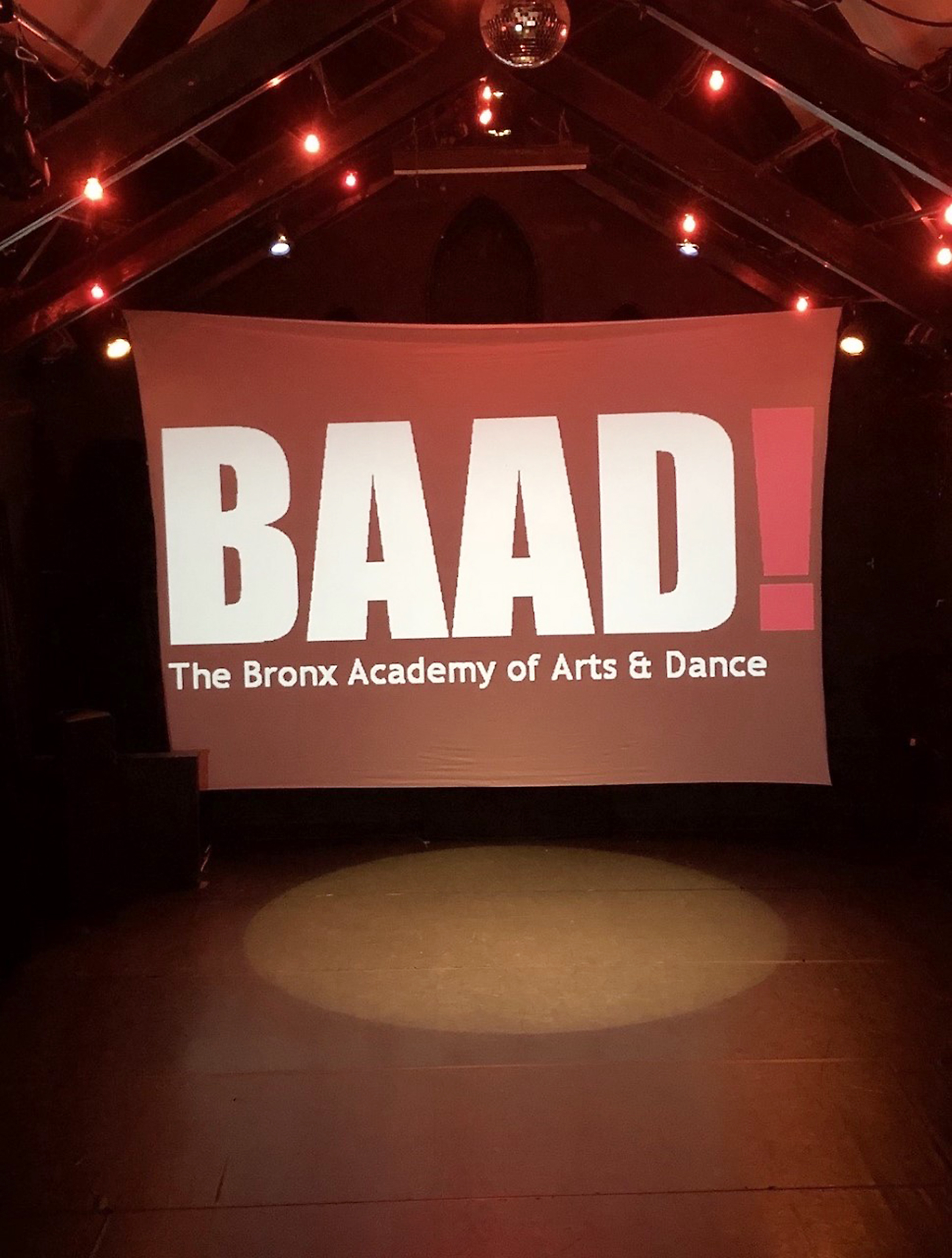 Red Banner with white writing that reads BAAD! The Bronx Academy of Arts & Dance hung on a stage with a spotlight shining down. Red Banner with white writing that reads BAAD! The Bronx Academy of Arts & Dance hung on a stage with a spotlight shining down.