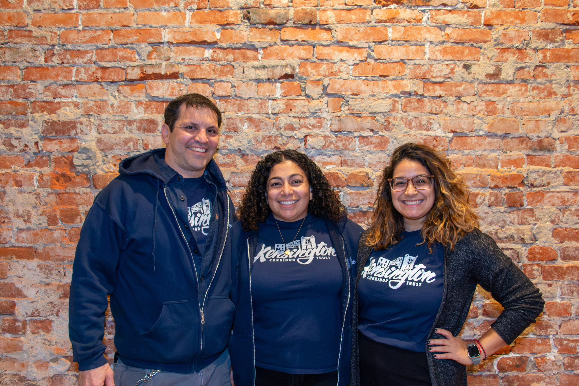 Three people stand in front of a brick wall, all wearing dark blue t-shirts with KCT's logo underneath dark hoodies.