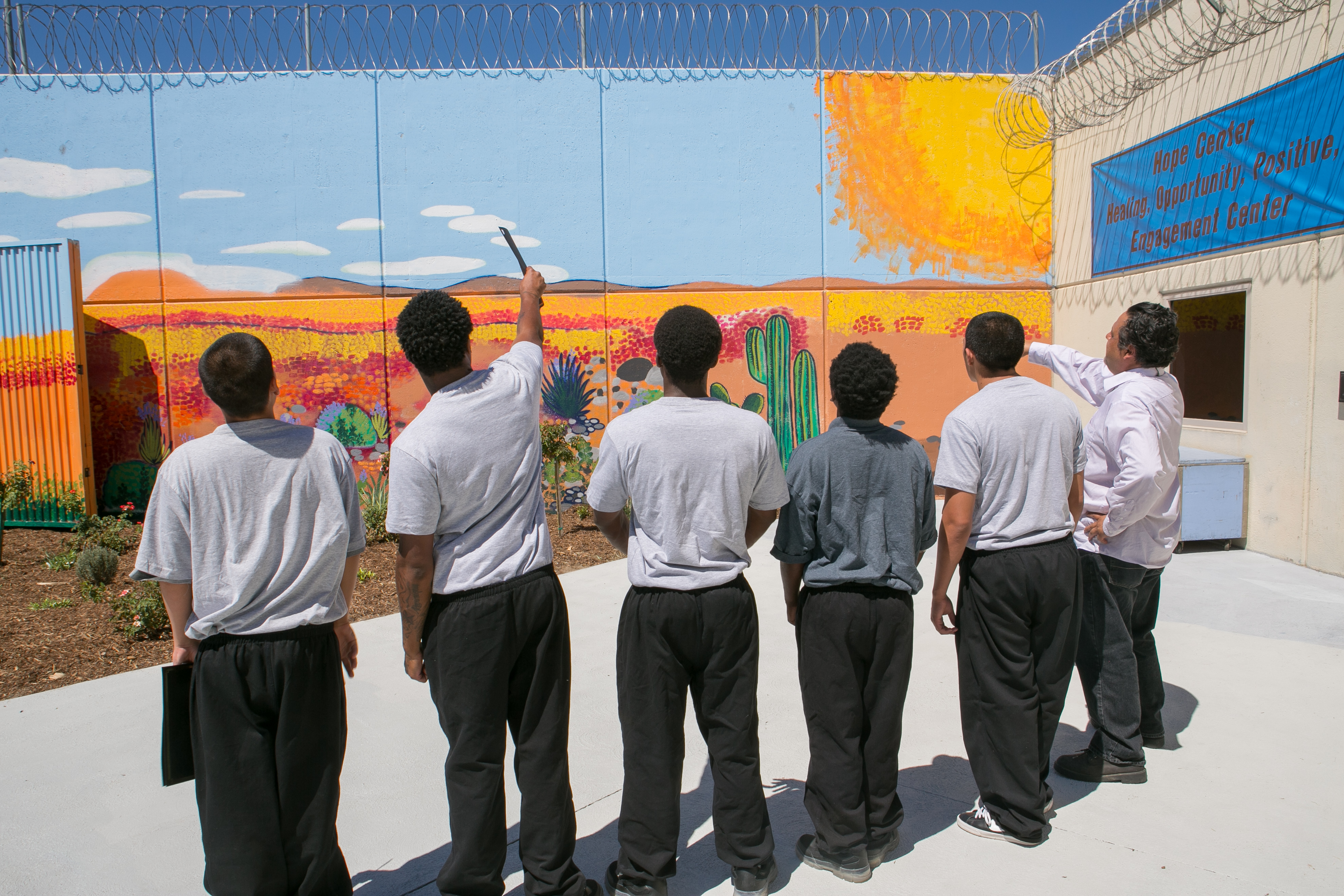 A group of six men stand facing a mural painted with golden fields and a sun shining to the right. Two of the men are pointing at different parts of the mural. A group of six men stand facing a mural painted with golden fields and a sun shining to the right. Two of the men are pointing at different parts of the mural.