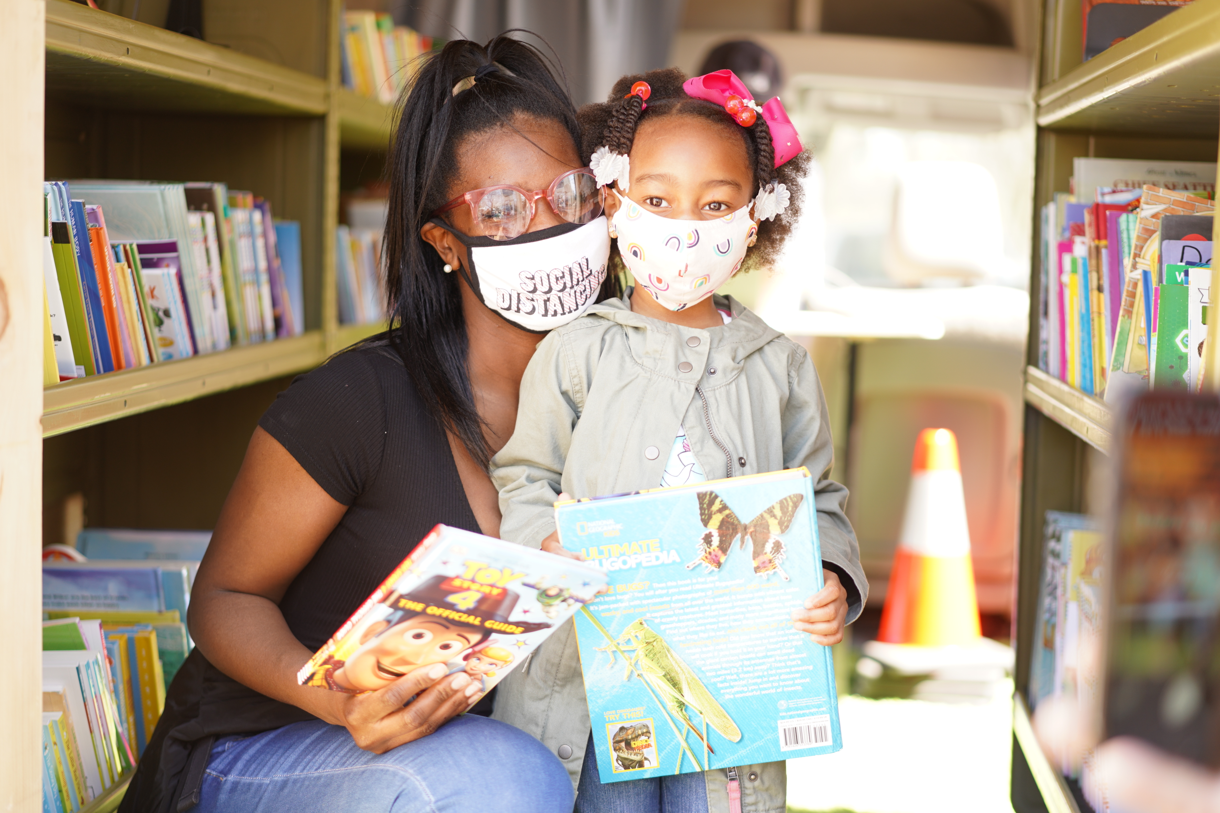A woman kneeling next to a child in between shelves of books, both are wearing masks and holding a children's book.