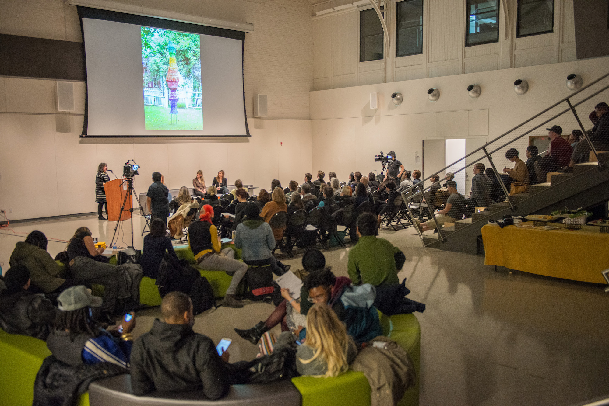 A photograph of a large crowd of people listening to artist Antonia Perez as she speaks about her artwork. Antonia stands to the far left at a podium in a large amphitheater with white walls; behind her, a massive projector screen shows an image of a rainbow hued woven sculpture entitled "Light Spectrum" that is made out of wire lampshade frames and crocheted plastic bags. Audience members fill the space, sitting on black folding chairs, green benches, or the amphitheater steps. A photograph of a large crowd of people listening to artist Antonia Perez as she speaks about her artwork. Antonia stands to the far left at a podium in a large amphitheater with white walls; behind her, a massive projector screen shows an image of a rainbow hued woven sculpture entitled "Light Spectrum" that is made out of wire lampshade frames and crocheted plastic bags. Audience members fill the space, sitting on black folding chairs, green benches, or the amphitheater steps.