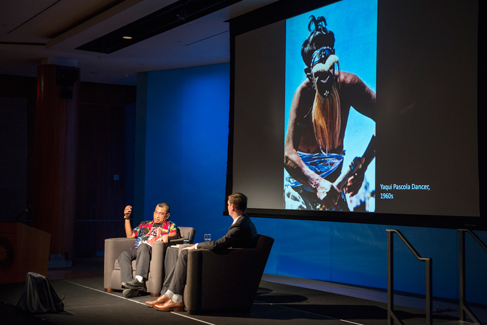 A photograph of two adults, Mario Martinez and Steven O'Banion, sitting in grey arm chairs on a raised platform in a darkened theater. Mario, who is seated on the left, is an older medium-skinned man wearing glasses and a colorful printed shirt. He looks downward at a monitor showing an image of a Native American Yaqui Pascola dancer in traditional garb that is also projected on a large screen behind him. Steven, on the right, is a light-skinned man in a dark grey suit, and has his back to the camera. A photograph of two adults, Mario Martinez and Steven O'Banion, sitting in grey arm chairs on a raised platform in a darkened theater. Mario, who is seated on the left, is an older medium-skinned man wearing glasses and a colorful printed shirt. He looks downward at a monitor showing an image of a Native American Yaqui Pascola dancer in traditional garb that is also projected on a large screen behind him. Steven, on the right, is a light-skinned man in a dark grey suit, and has his back to the camera.