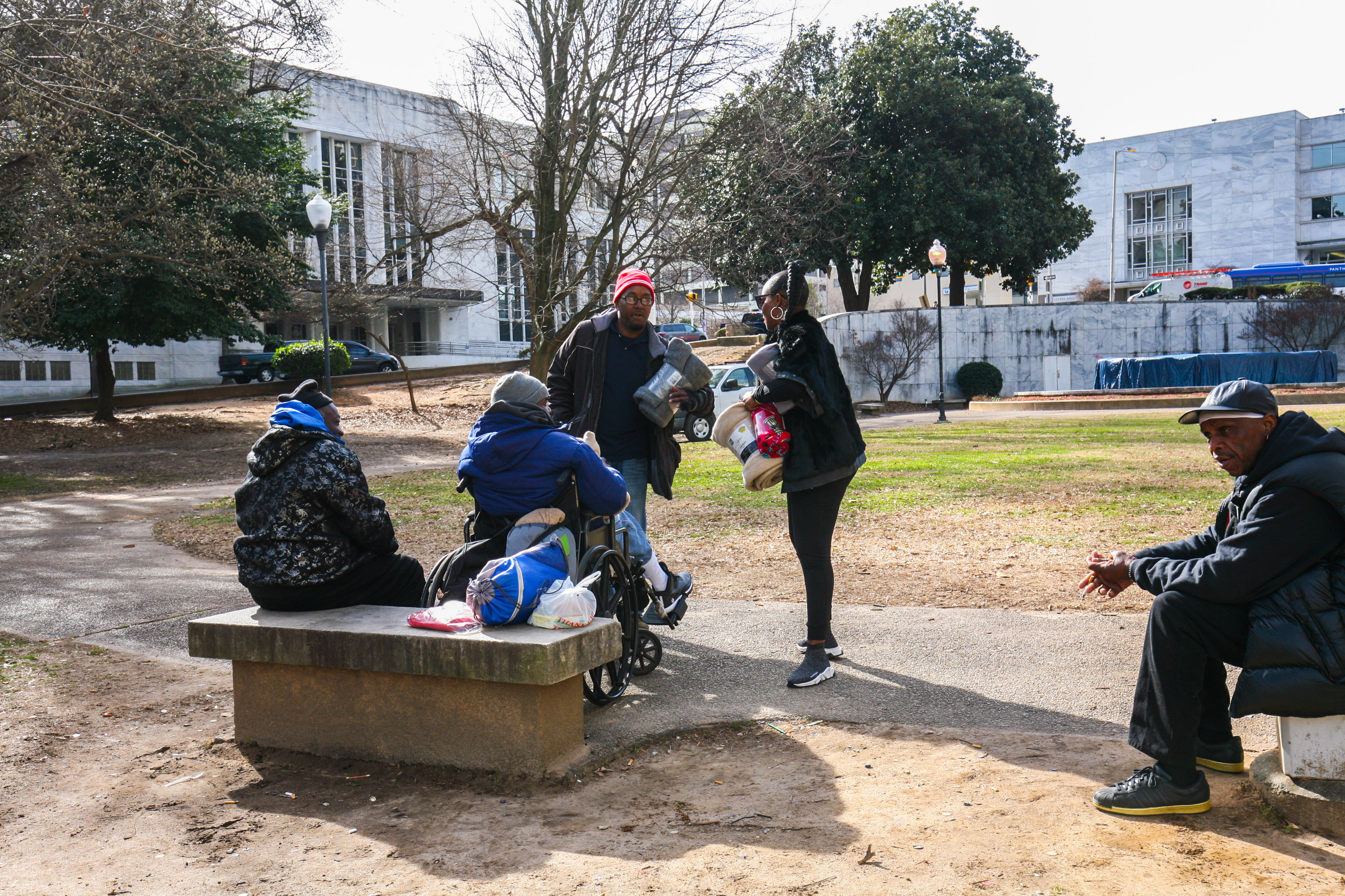 Kenya is meeting with a small group of people in a park to provide blankets; one person is in a wheelchair, and two others are seated on cement benches. Kenya is meeting with a small group of people in a park to provide blankets; one person is in a wheelchair, and two others are seated on cement benches.
