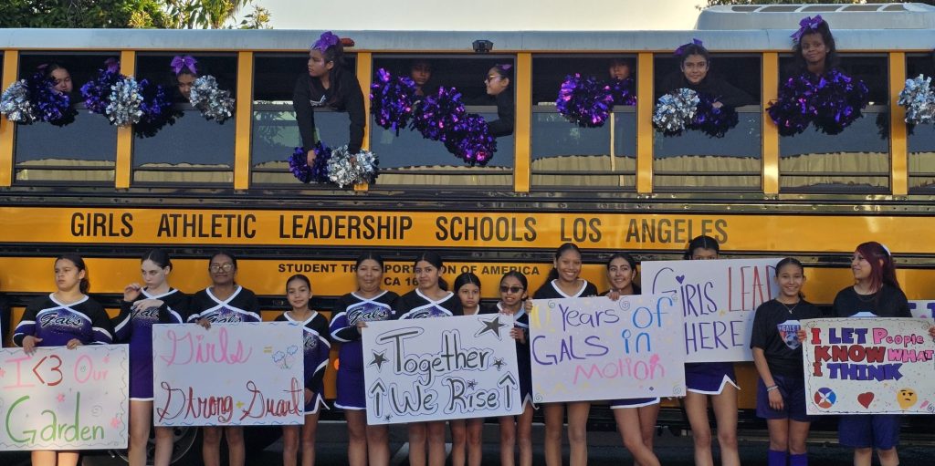 “A group of smiling students hold up signs while standing in front of a GALS LA school bus.