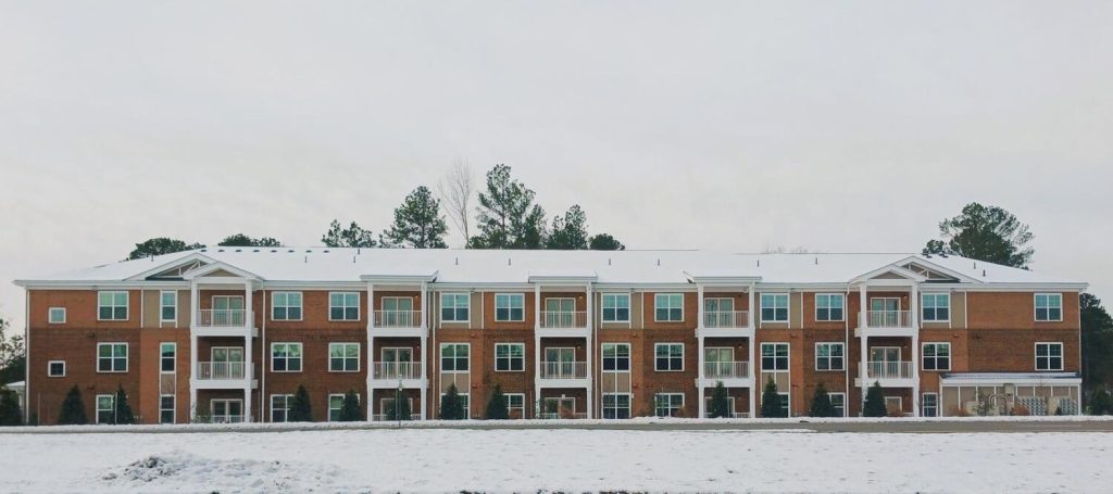 A multi-story brick building with white window frames and snow on the roof and ground in front.