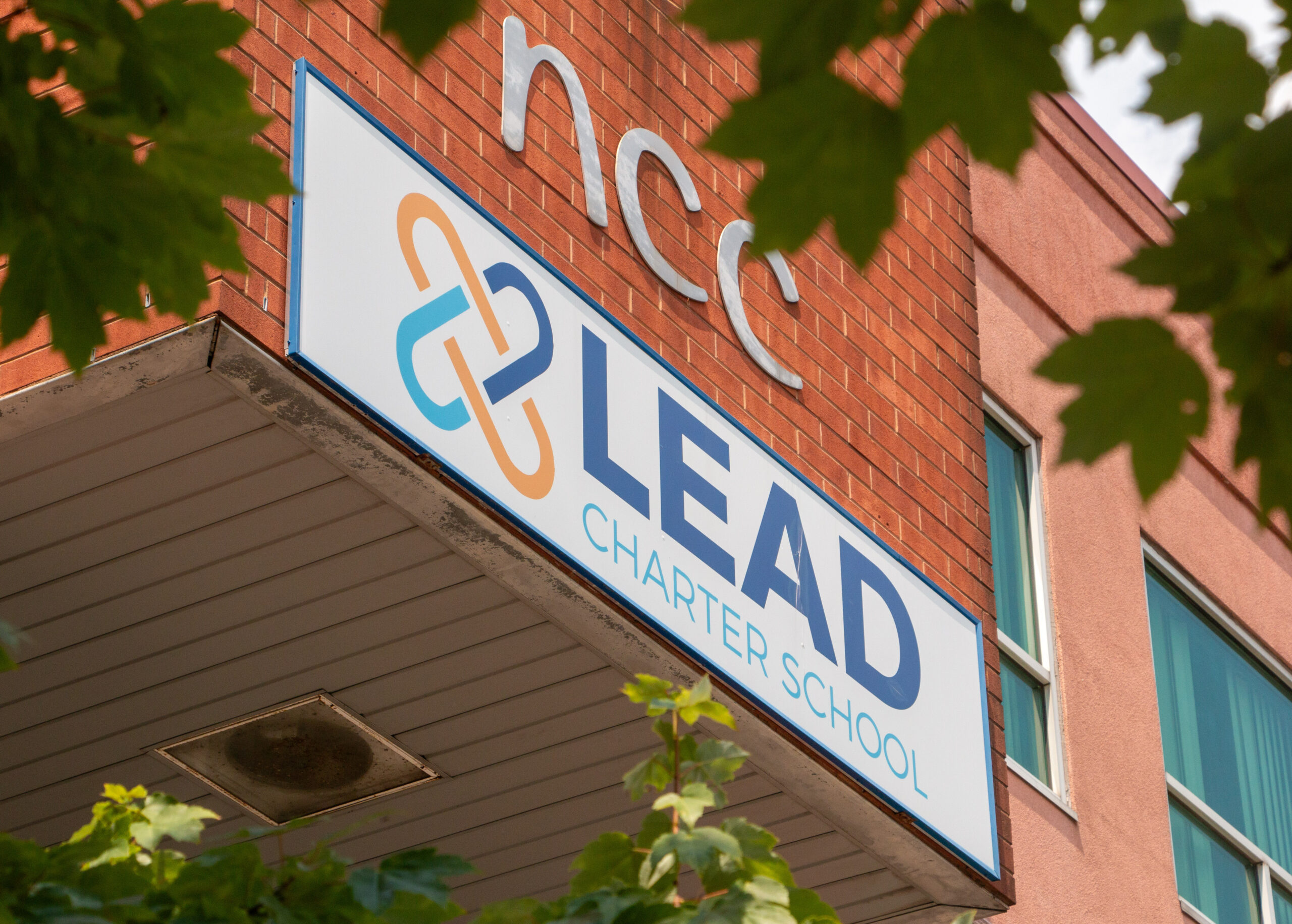 Sign for Lead Charter School on a brick building, partially obscured by green leaves. The sign features a logo and the text is clear and bold. Sign for Lead Charter School on a brick building, partially obscured by green leaves. The sign features a logo and the text is clear and bold.