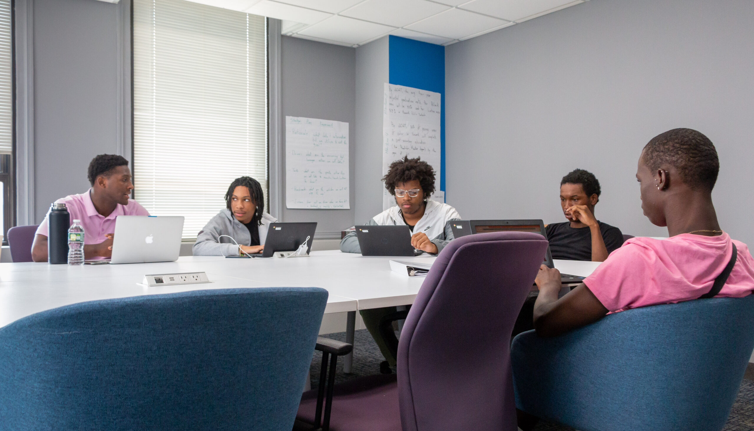 Five people sit around a conference table with laptops, engaged in discussion. The room has a focused, collaborative atmosphere. Whiteboards are in the background. Five people sit around a conference table with laptops, engaged in discussion. The room has a focused, collaborative atmosphere. Whiteboards are in the background.