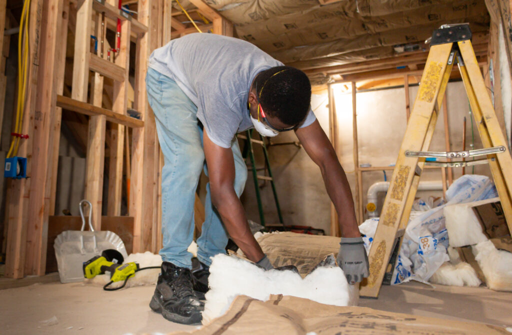 A person in protective goggles and mask installs insulation in a partially constructed room, conveying focus and productivity. A ladder and tools are nearby. A person in protective goggles and mask installs insulation in a partially constructed room, conveying focus and productivity. A ladder and tools are nearby.