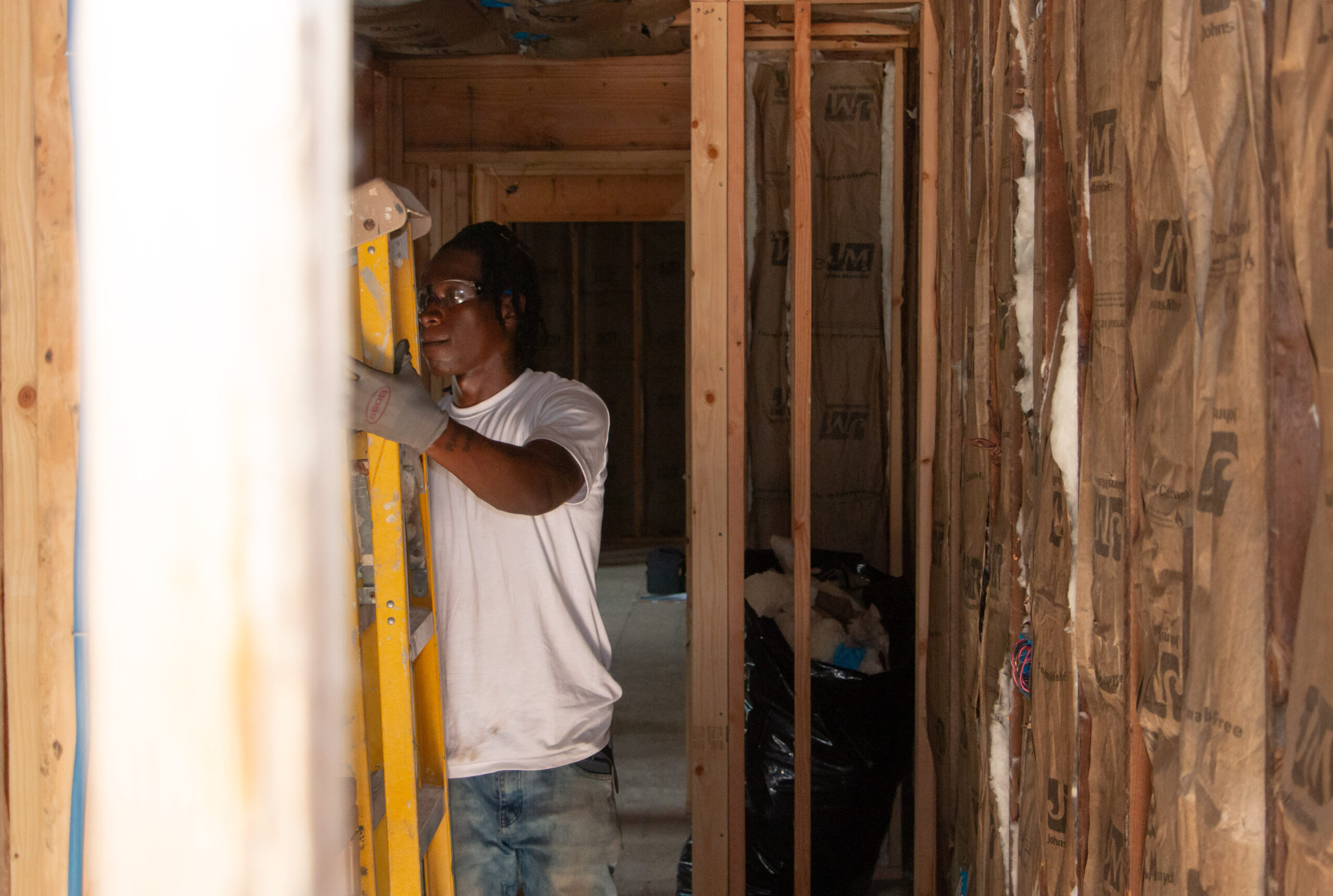 A person wearing a white shirt and safety glasses stands on a yellow ladder in an unfinished room with exposed wooden beams and insulation. The scene conveys concentration and construction work in progress. A person wearing a white shirt and safety glasses stands on a yellow ladder in an unfinished room with exposed wooden beams and insulation. The scene conveys concentration and construction work in progress.