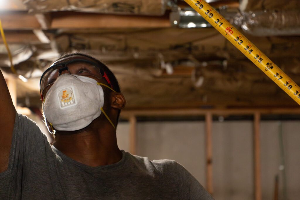 Man in a construction setting wears a mask and goggles, holding a tape measure against an unfinished ceiling, focused and attentive to detail. Man in a construction setting wears a mask and goggles, holding a tape measure against an unfinished ceiling, focused and attentive to detail.