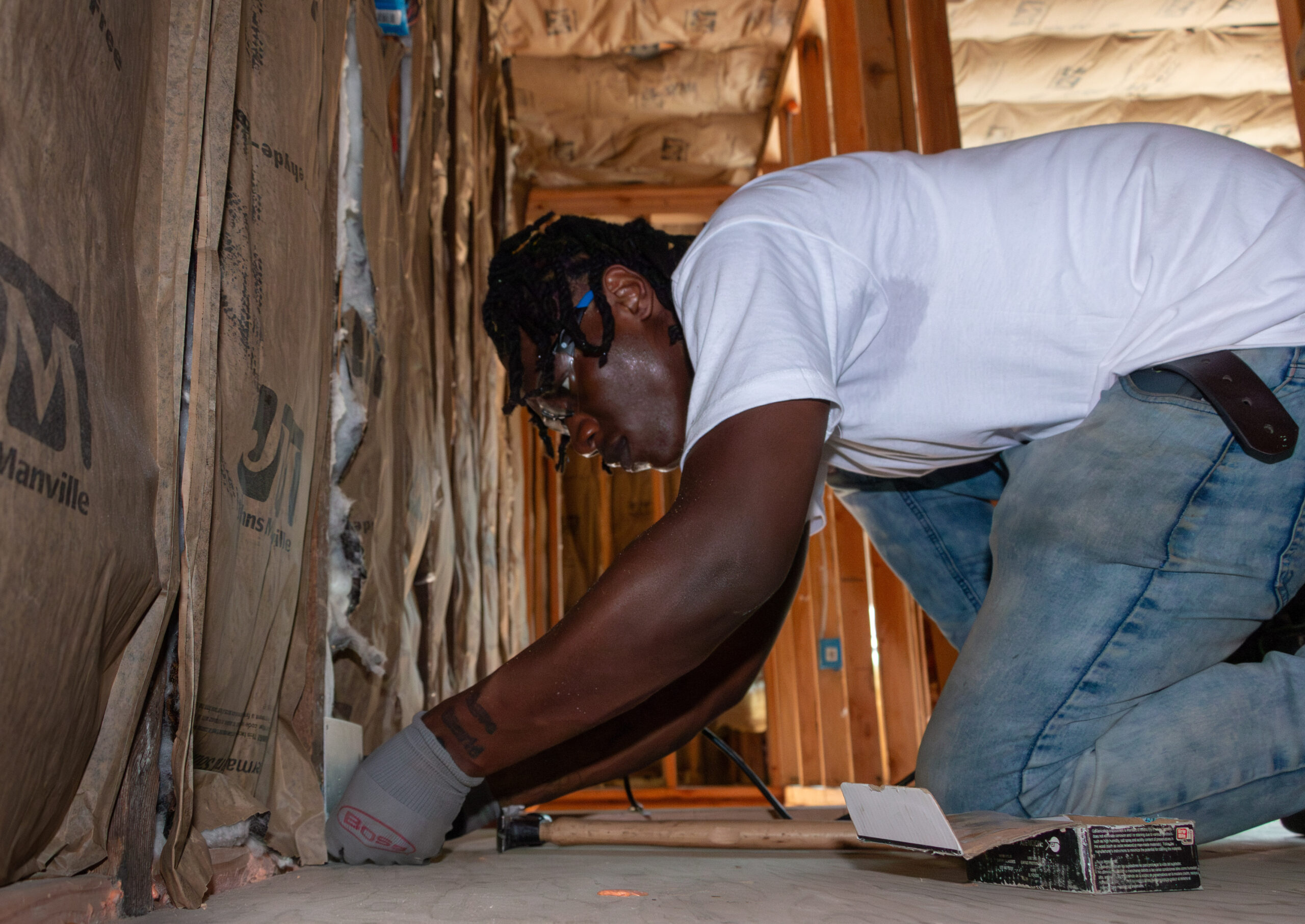 A person wearing a white T-shirt and jeans is kneeling, installing insulation in a wooden-framed room. They appear focused and diligent. Tools are nearby. A person wearing a white T-shirt and jeans is kneeling, installing insulation in a wooden-framed room. They appear focused and diligent. Tools are nearby.