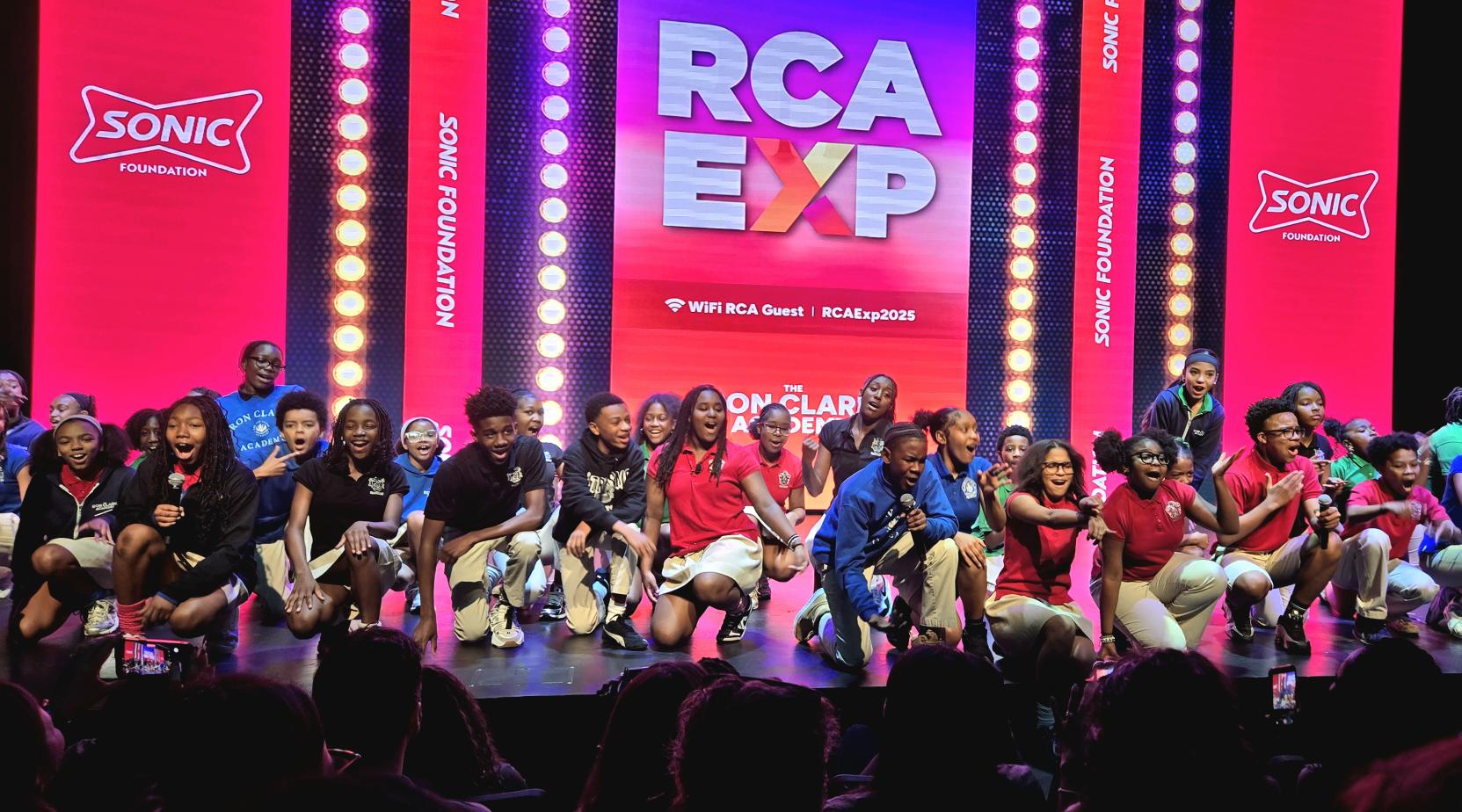 RCA students kneeling onstage in front of a bright pink and red backdrop as part of a welcome performance for NFF’s staff site.