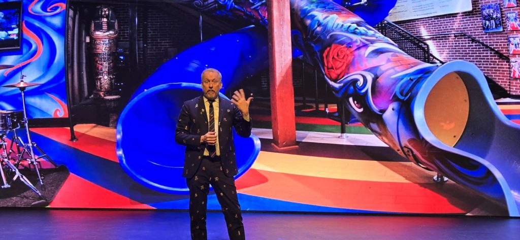 Ron Clark speaking into a microphone as he speaks about RCA in front of a colorful image of school slides.