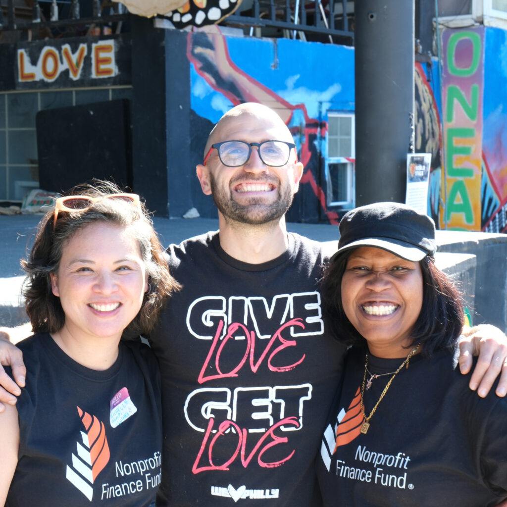 Three smiling colleagues—a man between two women—pose with their arms around each other in front of a colorful outdoor mural. Two wear shirts for the "Nonprofit Finance Fund," while the man's shirt reads, "GIVE LOVE GET LOVE."